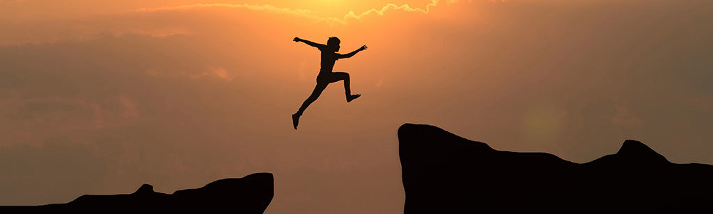 Eine Person springt vor rötlichem Himmel über eine Klippe